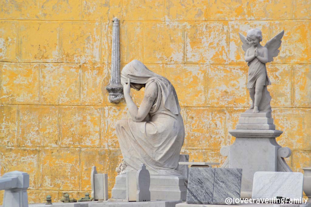 A seated mourning figure and an angel beside stone memorials against a weathered yellow wall at Colón Cemetery.
