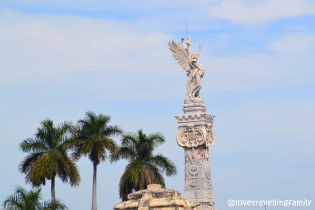 A sculpted figure above an ornate stone memorial, seen against palms and blue sky in Colón Cemetery.