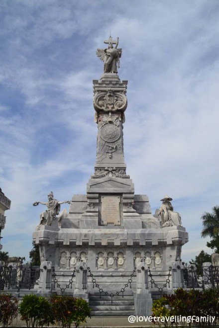 The tall firefighters’ monument at Colón Cemetery, with carved figures, an iron enclosure and flowers.