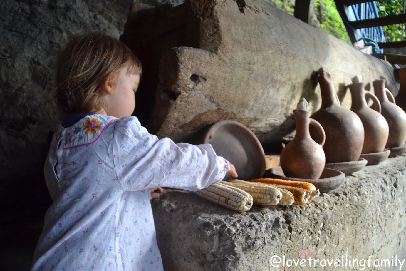 A toddler in white explores old clay jugs and corn on a stone ledge at a guesthouse in Kutaisi.
