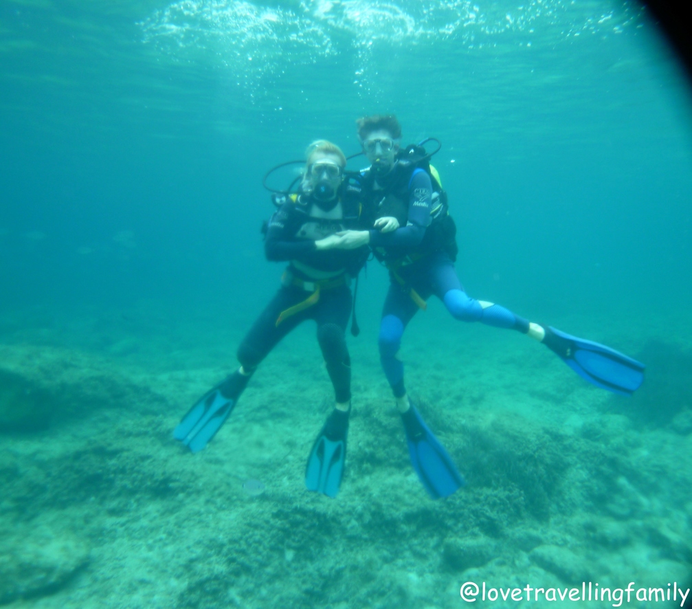 Two divers in wetsuits pose underwater in clear turquoise water off Cala Rajada, Mallorca.