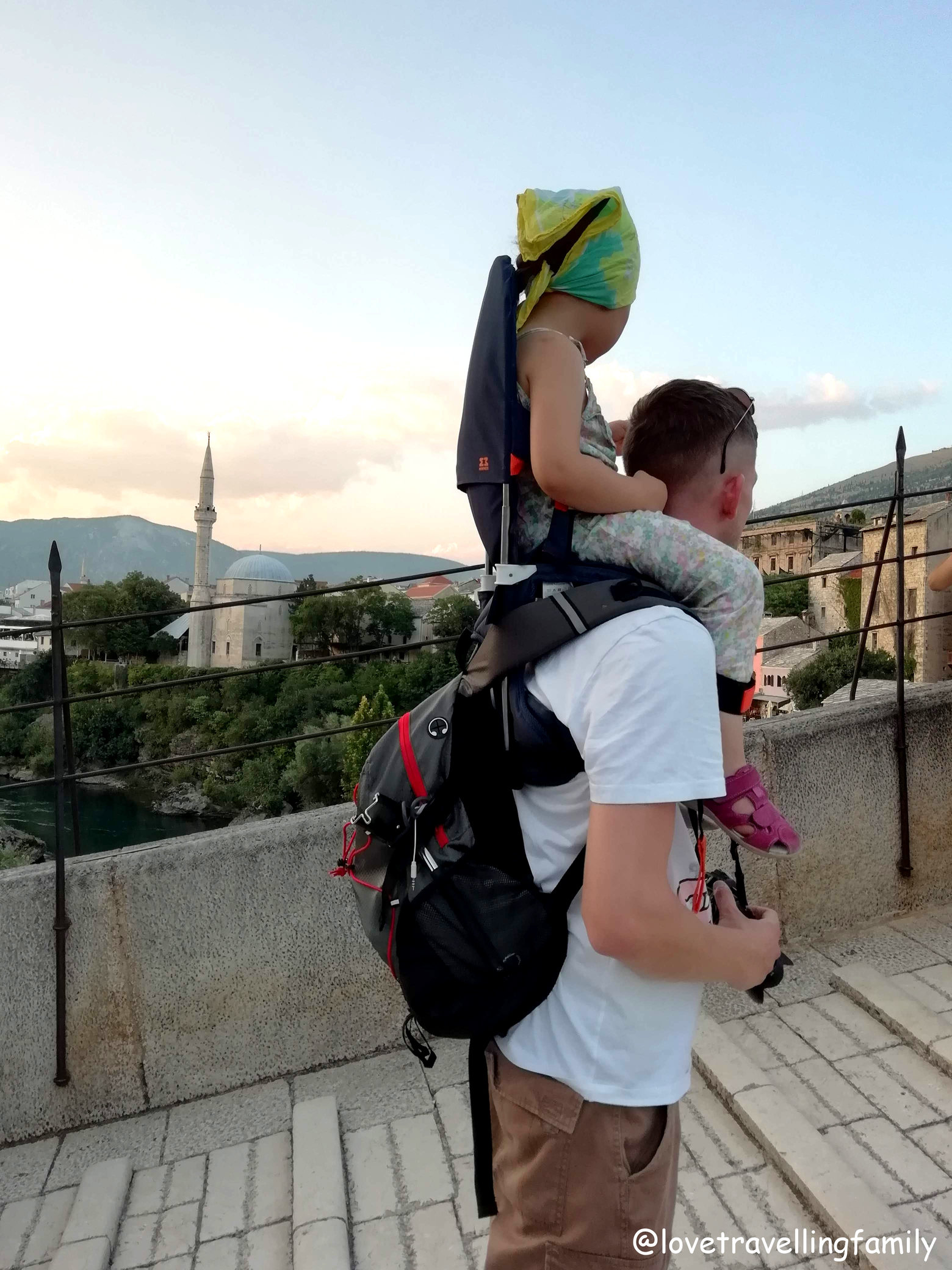 A parent carrying a young child in an older MiniMeis shoulder carrier during the family’s journey.