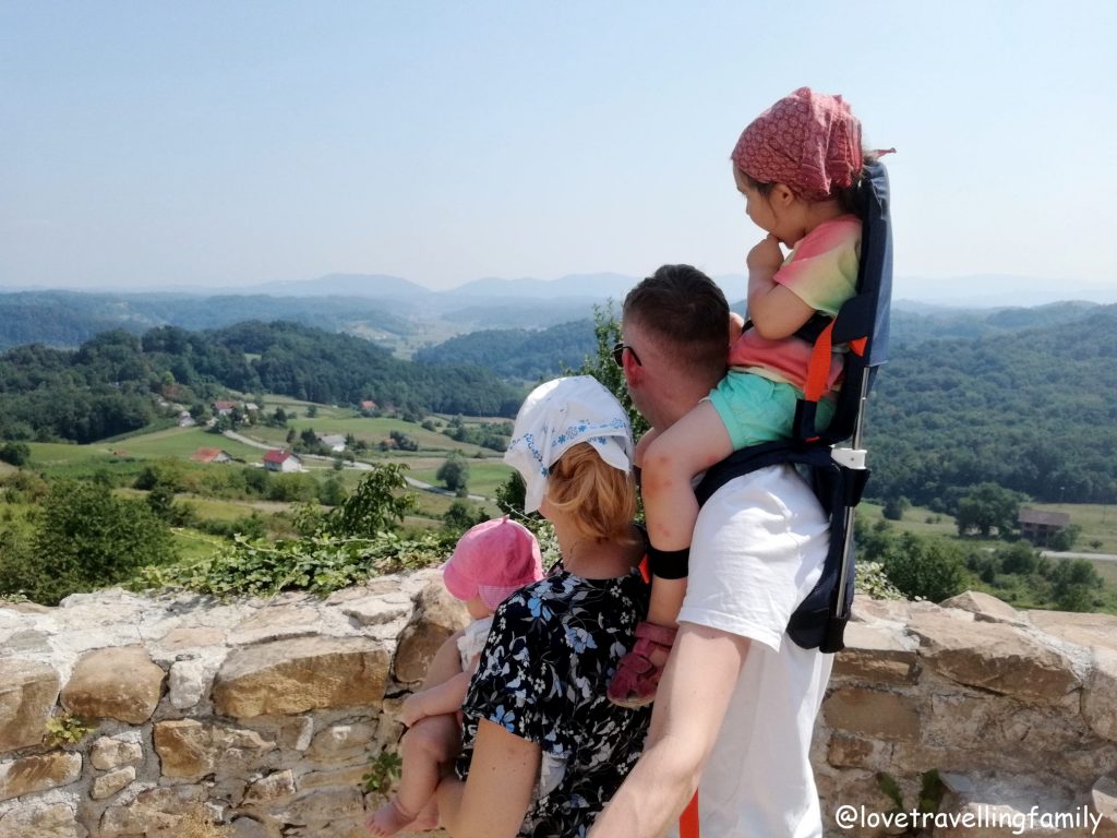 A parent and children beside a stone parapet overlooking a green landscape, with one child in a shoulder carrier.