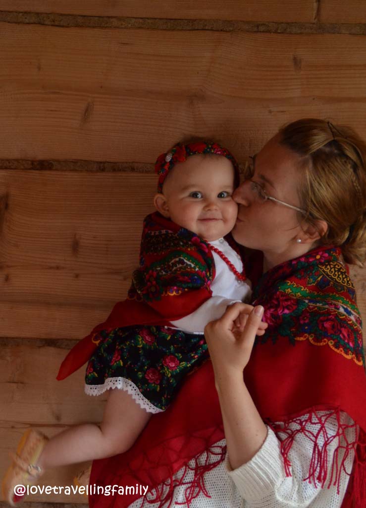 A mother kisses her baby, both wearing floral clothing, beside a timber wall in Podhale.