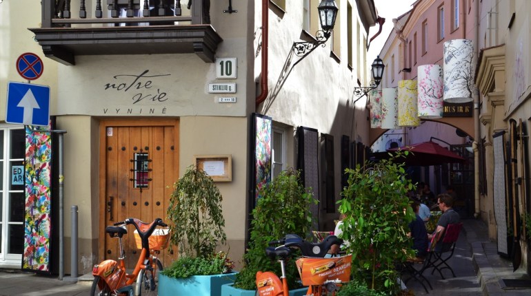 A narrow old-town street in Vilnius with a café doorway, flower pots and bicycles.